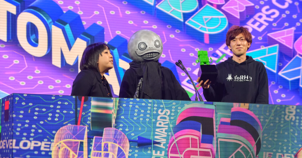 Yoko Taro con la maschera di Emil affiancato da due persone sul palco dei Game Developers Choice Awards